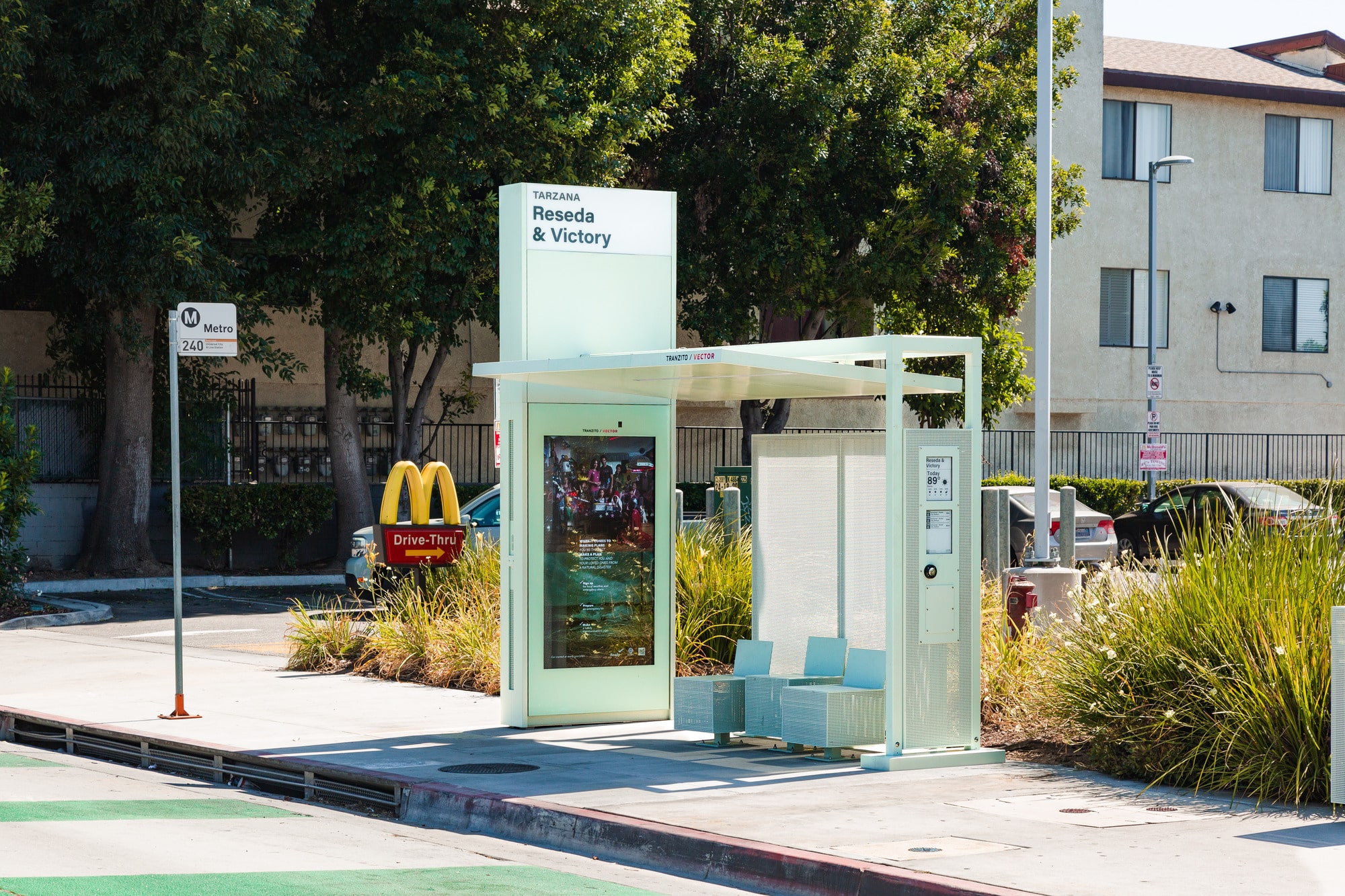A bus shelter in Tarzana is seen from the front right with three seats, a digital display on the left side and rider information displayed on the right side panel.
