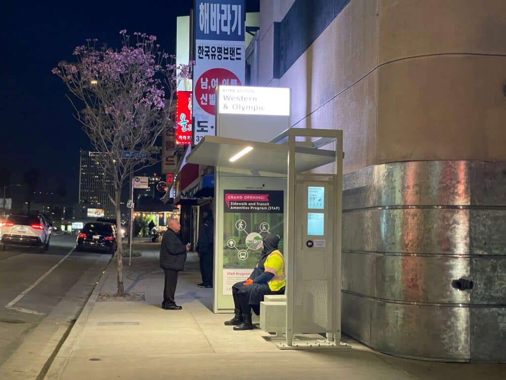 A bus shelter in Koreatown is seen at night with two riders awaiting the next bus in a well lit shelter.