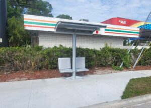 A small bus shelter is seen along a sidewalk with a single post, an awning over head and one seat on each side of the shelter post.