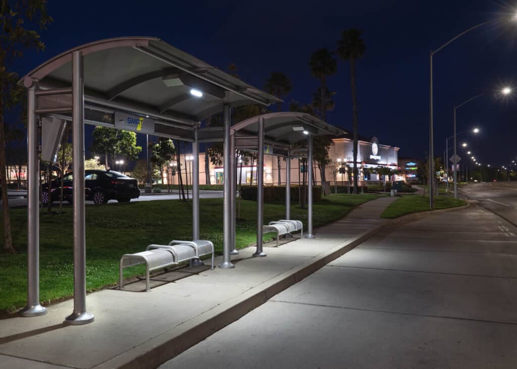 Two bus shelters are seen side by side at night from the left side front with bench seating for three under both and lighting.