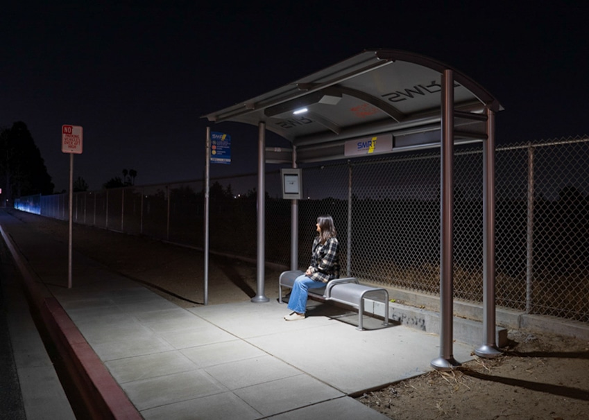A rider is seen waiting at night under a lit bus shelter with rider information display on a back left screen and bench seating for three.