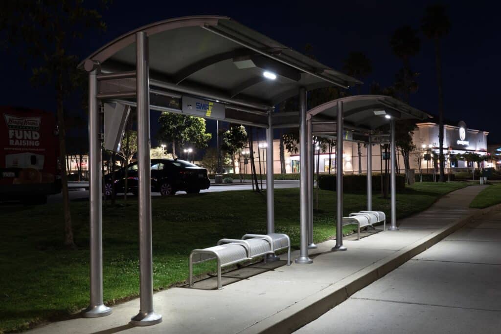 Two bus shelters are seen from the left side at night with ample lighting for each shelter and its bench seating.