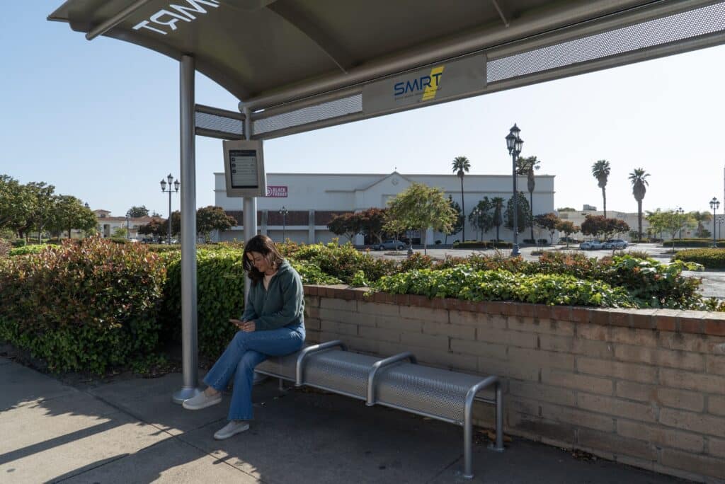 A woman is sitting on a bench under a bus shelter looking at her phone awaiting the next bus.