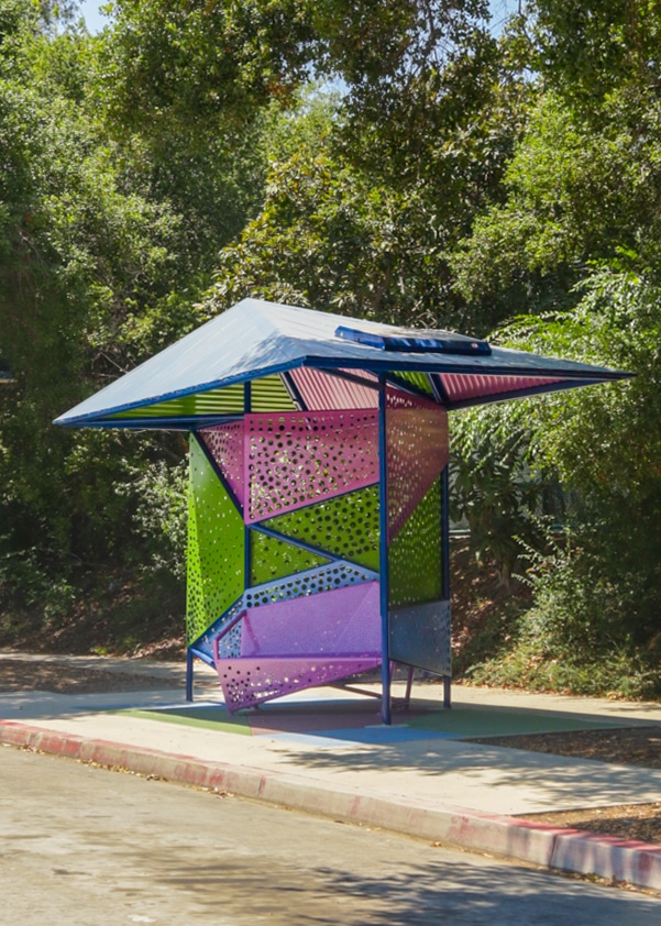 A colorful bus shelter is seen from the right side with a purple bench, blue roof and multicolored back panels with a hole design.