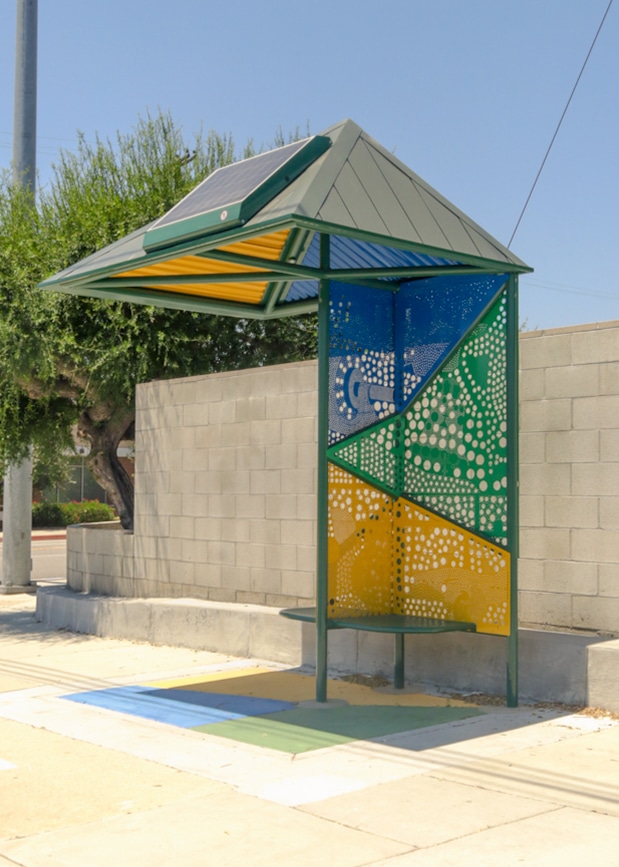 A bus shelter is seen with a solar paneled roof, hole back panel designs and seating for two all in a colorful green, yellow and blue.
