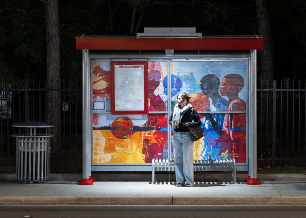 A bus shelter with a rider information display on the back panel and a rider waiting for the bus under the shelter at night with ample lighting.