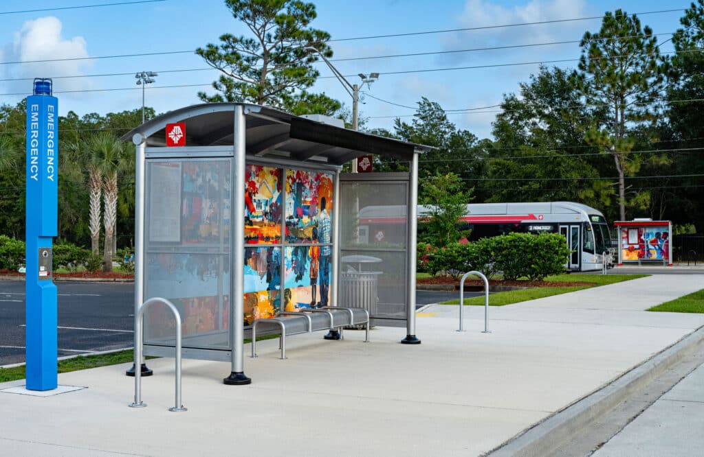 A custom art bus shelter is seen from the left front with bike stands on either side of the shelter and bench seating for four under the shelter.