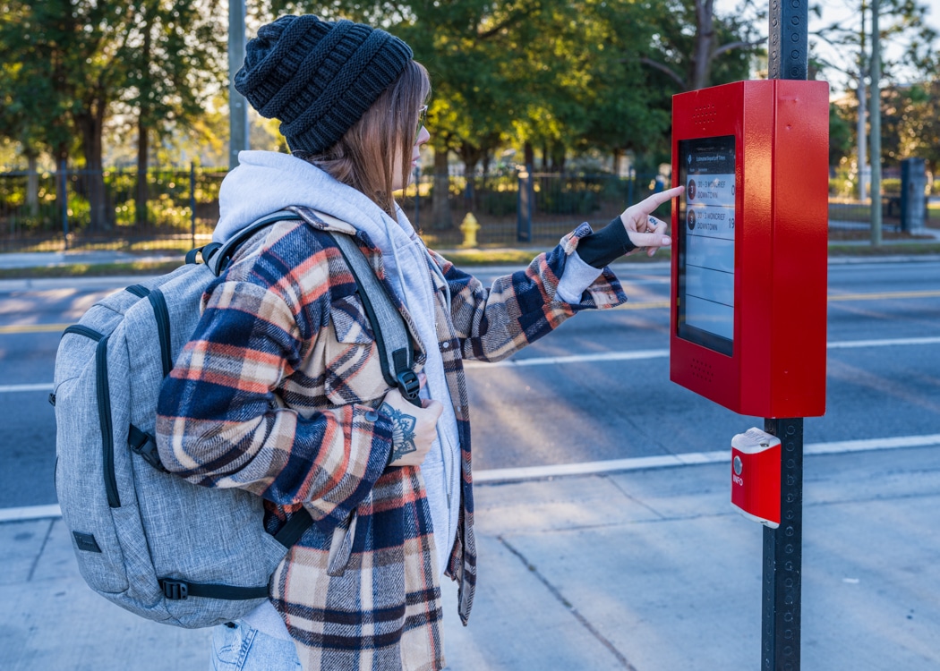 A rider is pointing at the rider information display located on a transit pole with an info button below the display.