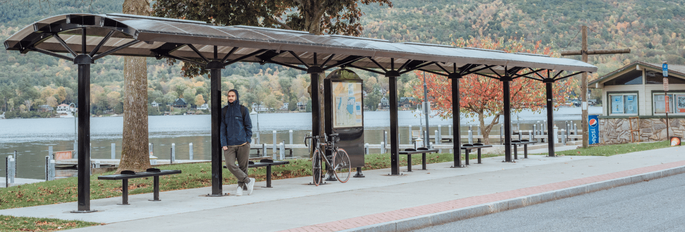 A man is leaning against on of the posts of a bus shelter with multiple benches under the long shelter and a rider information display in the middle.